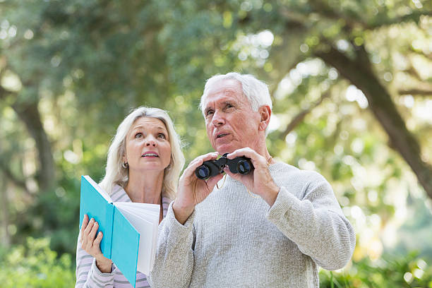 Senior couple outdoors in the woods or a park with binoculars, bird watching.  The woman is holding a book open, trying to identify the types of birds they are seeing.  They are both looking upward, talking.  There are trees in the background.